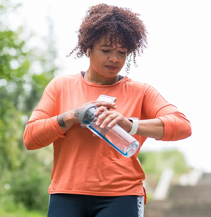 Person outdoors in an orange shirt checks a smartwatch while holding a water bottle.