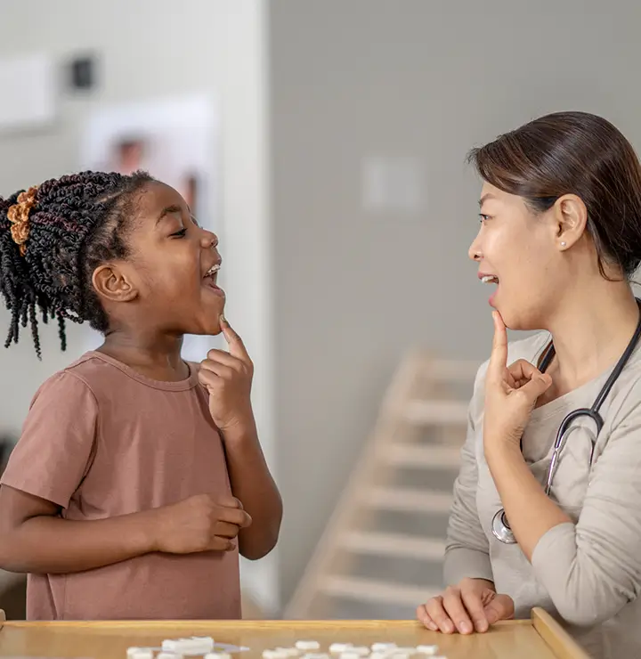 A child and a healthcare professional practice speech exercises, smiling at a friendly table setup.