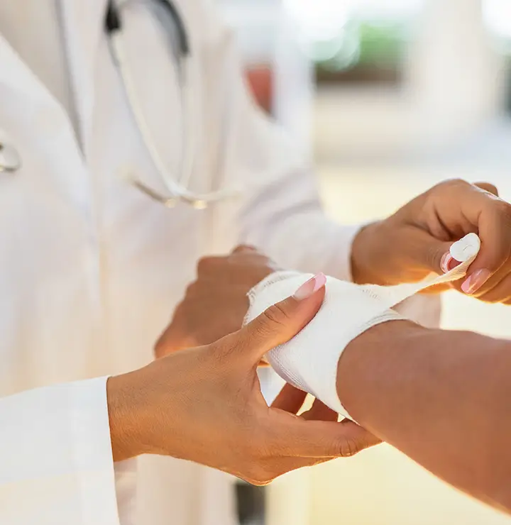 A person in a white coat wraps another’s wrist with a bandage in a softly blurred setting.