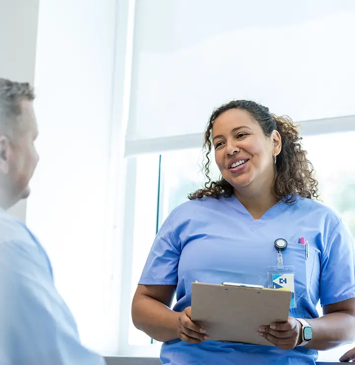 A healthcare worker in scrubs holds a clipboard, smiling while speaking to a seated person.