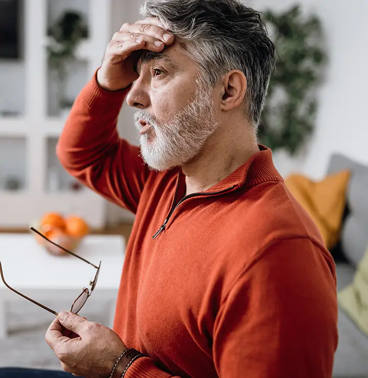 Concerned man holding glasses and touching his forehead, sitting on a couch.