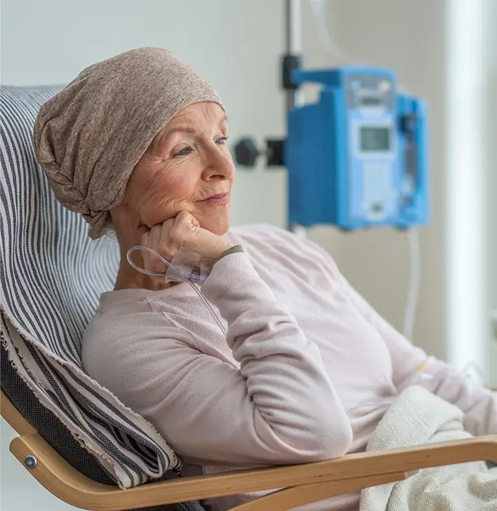 An older woman in a headscarf receives IV treatment, resting her chin on her hand thoughtfully.