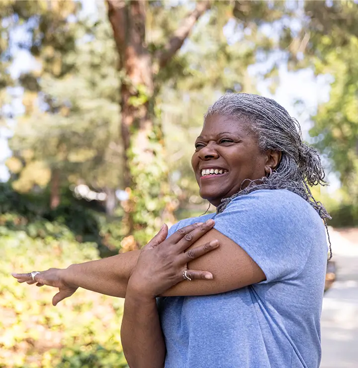A joyful woman in a blue shirt stretches her arms outdoors on a sunny day surrounded by greenery.