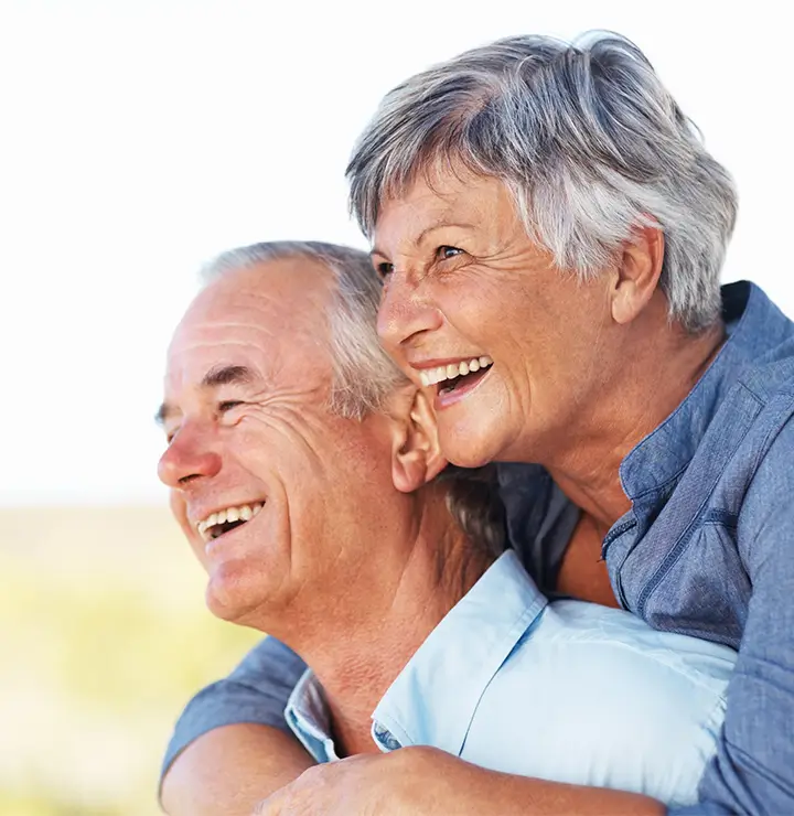 An elderly couple smiles warmly, the man in the foreground and the woman embracing him from behind.