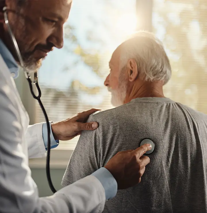 A doctor listens to an older man’s chest with a stethoscope as sunlight filters through a window.