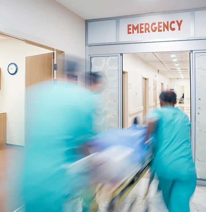 Healthcare professionals in blue scrubs rush a patient on a stretcher through emergency doors. The scene reflects urgency, with movement in the hospital hallway.