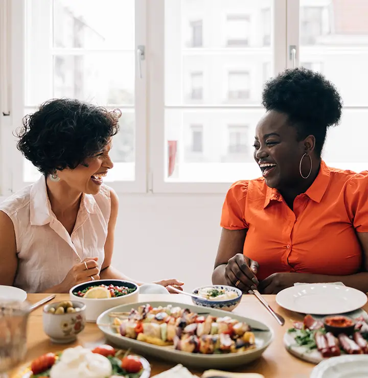 Two women sit at a table, smiling and chatting while enjoying colorful dishes. The room is bright, with large windows letting in natural light.