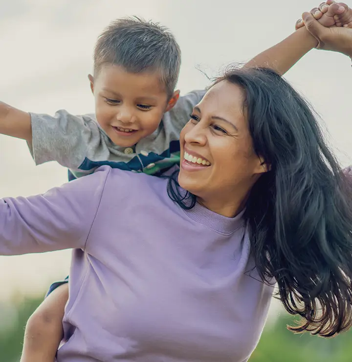 A joyful woman carries a smiling young boy on her back. He stretches his arms wide playfully while the sky forms a cheerful backdrop.