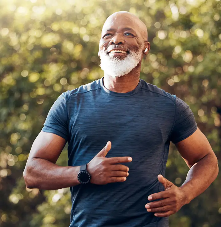 A bearded man jogs outdoors in a dark blue T-shirt, wearing a smartwatch and earbuds. He smiles under sunny skies, with trees in the background.