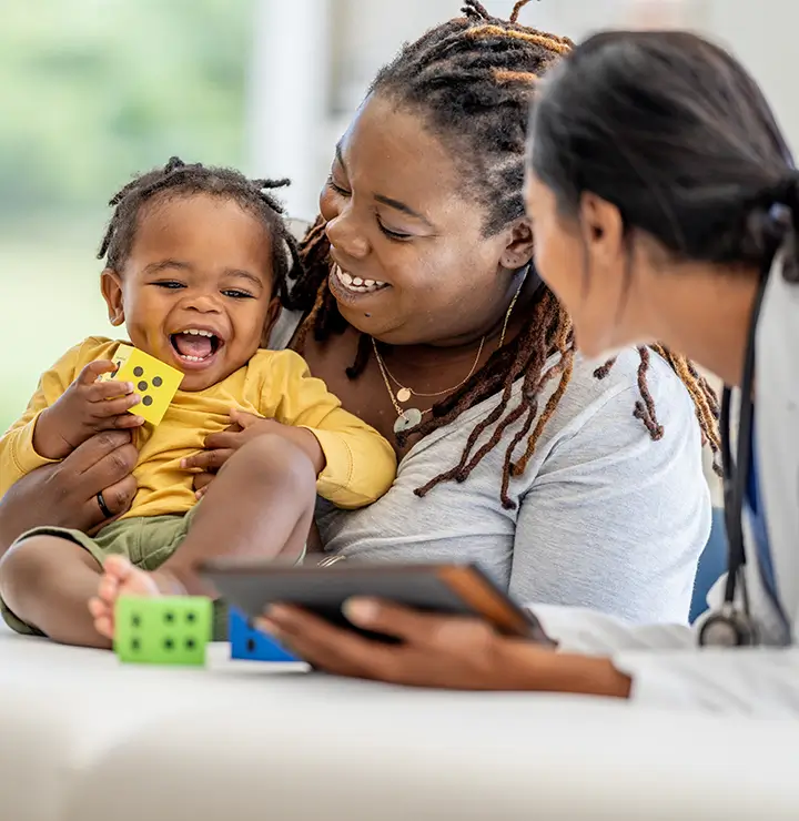 A smiling adult holds a laughing baby playing with toy blocks while a healthcare worker with a tablet engages with them in a medical setting.