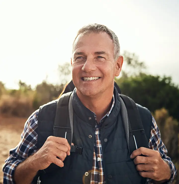 A smiling man with short gray hair, wearing a plaid shirt and vest, backpacks through a sunny, natural landscape in the background.