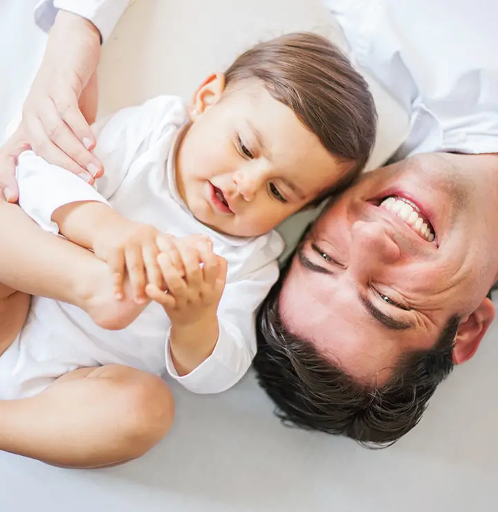 A man lies on his back, smiling broadly as a baby beside him holds its foot. They share a playful, joyful moment together.