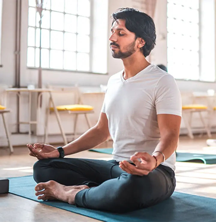 Person meditates cross-legged on a yoga mat in a sunny room with large windows.