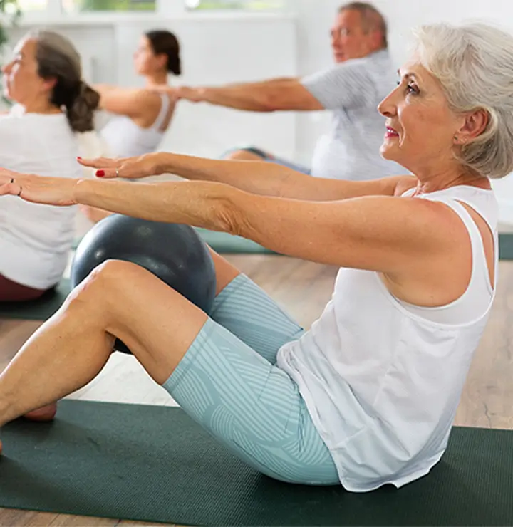 Elderly group exercises with yoga mats and balls in a bright room with wooden floors.