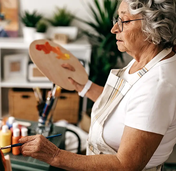 Elderly woman painting on a canvas in a cozy art studio, holding a palette with paint.