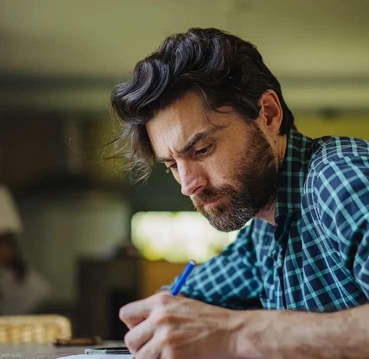 Man with a beard in a checkered shirt writing at a table with a focused expression, indoors with a blurred background.