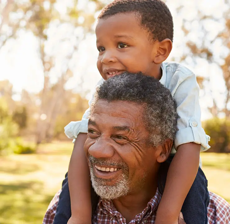 A smiling man carries a child on his shoulders in a sunny park setting.