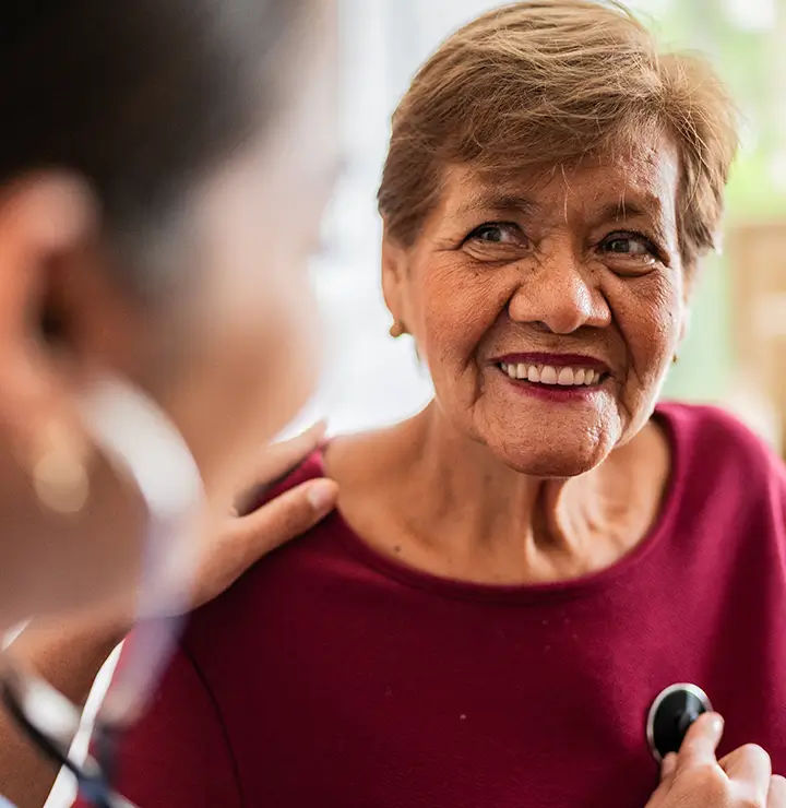 A healthcare professional uses a stethoscope on a smiling elderly person in a red shirt.