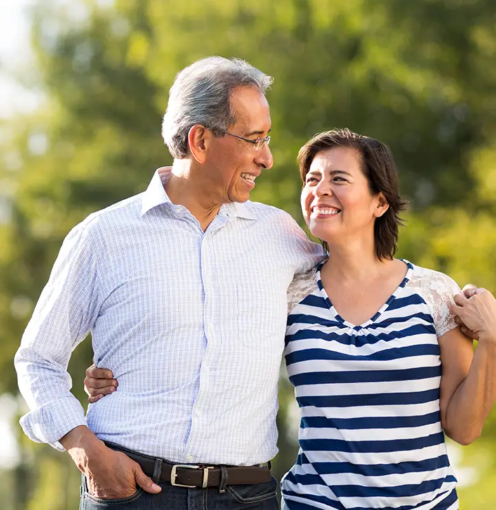 A smiling couple walks outdoors in a park, with trees in the background. The man wears a white shirt, and the woman is in a striped top.