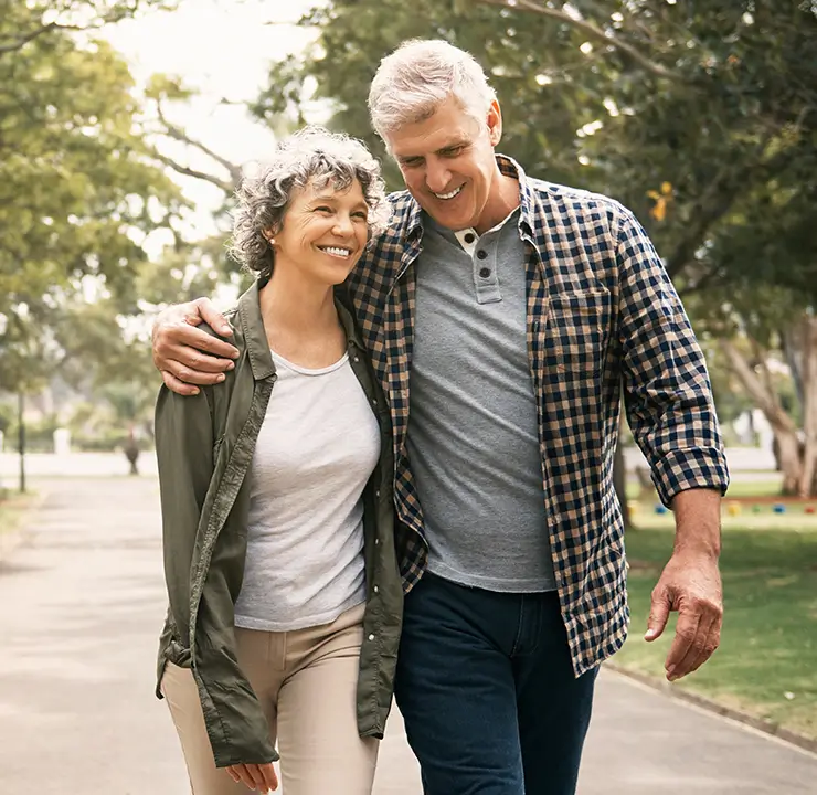 A smiling couple walks arm in arm through a park, surrounded by trees and greenery.