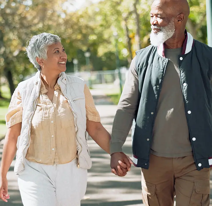 An elderly couple walks hand in hand through a sunny park, smiling at each other, surrounded by trees.