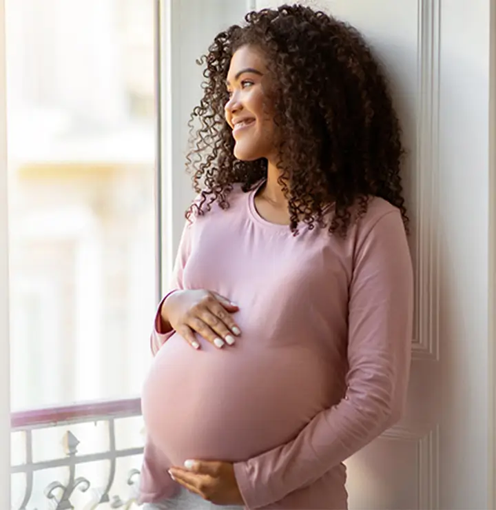 Pregnant woman smiles, holding her belly near a window with soft natural light.