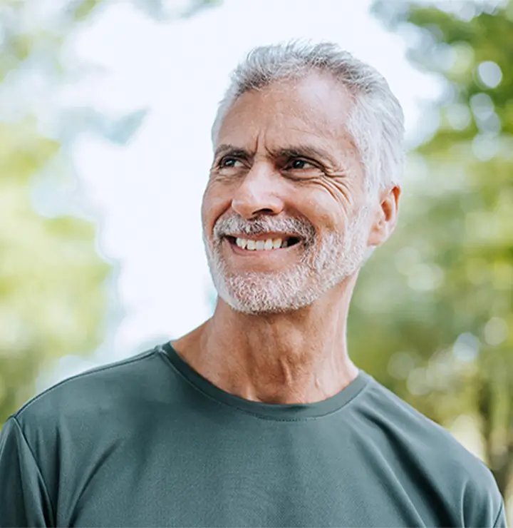 Smiling older man in a green shirt stands outdoors with trees blurred behind.