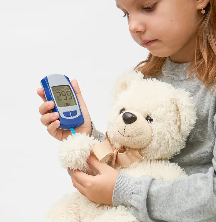 Girl uses a glucose meter while holding a teddy bear wearing a gray sweater.