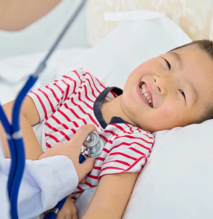 Child smiles in bed as a healthcare worker listens to their chest with a stethoscope.
