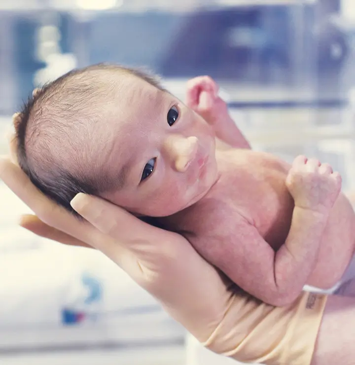 Healthcare worker holds a newborn baby in gloved hands in a hospital room.