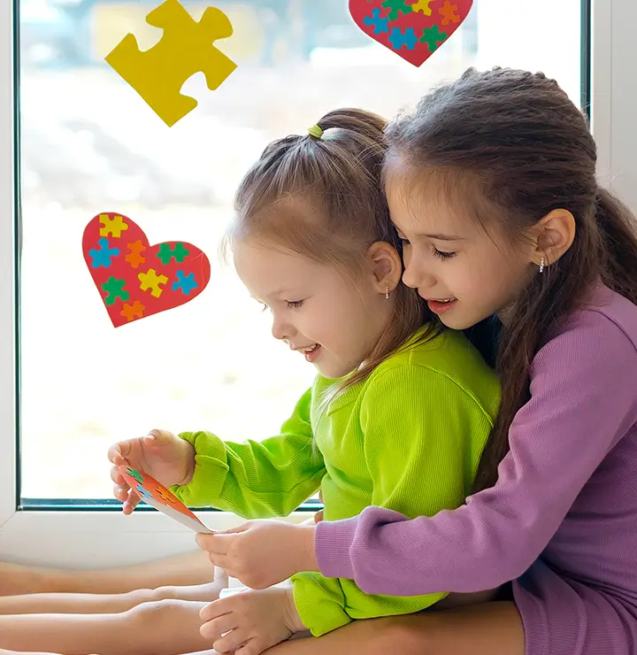 Two girls by a window with puzzles and hearts; one in purple hugs the other in green as they smile, holding a card.