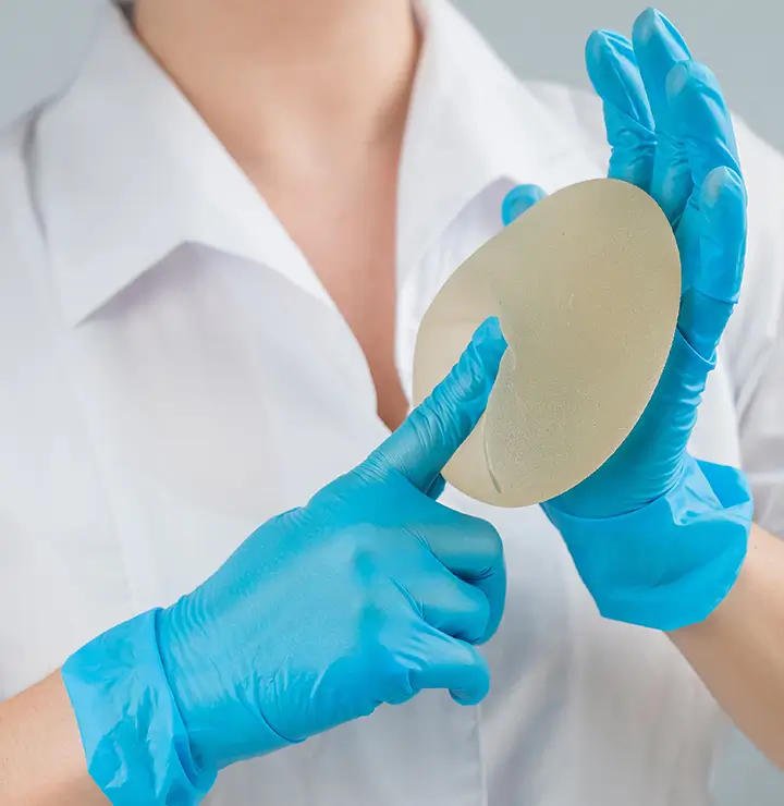 A gloved person in a white coat examines a silicone breast implant, pressing a finger into its surface against a neutral background.
