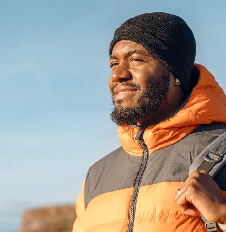 A person in a black hat and orange-gray jacket smiles, holding a backpack strap; sunlight warms their face against a clear sky.