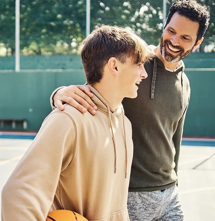 A man and teen smile on a basketball court; the teen holds a basketball, and the man rests a hand on their shoulder under a clear sky.