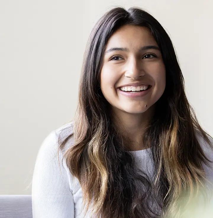 A person with long brown hair and a white shirt smiles softly against a light-colored background.