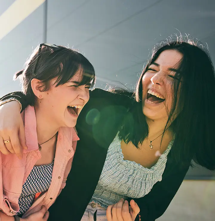 Two women laugh joyfully; one in a pink shirt and sunglasses, the other in a black cardigan. Sunlight flares near a modern building.