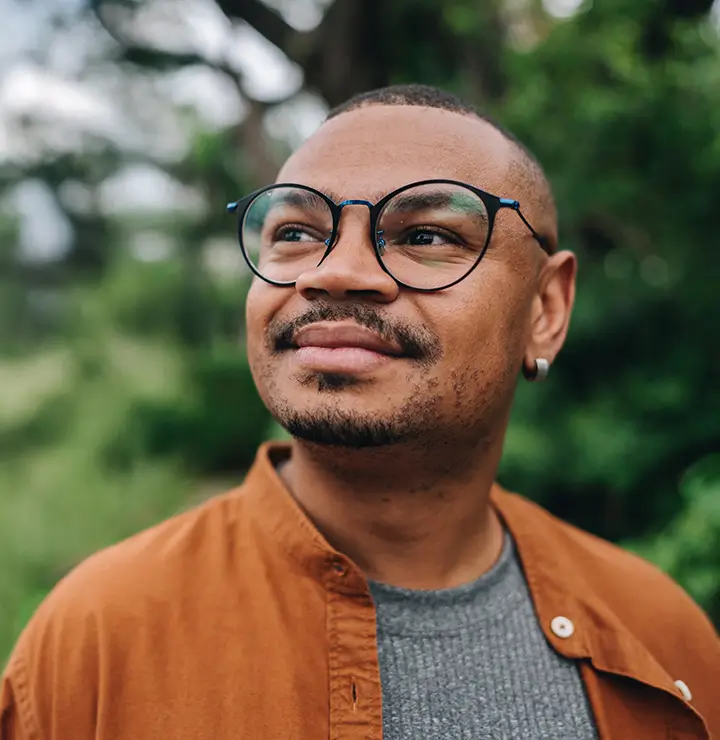 A person with glasses and a short haircut smiles while looking into the distance; blurred green foliage creates a serene backdrop.