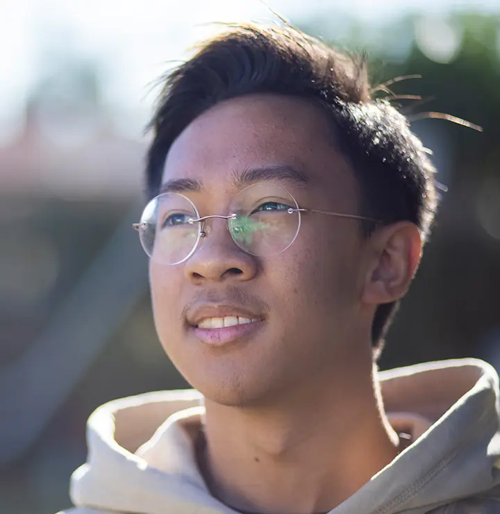 A person in glasses and a hoodie gazes upward with a calm look; soft greenery and sunlight blur in the background.