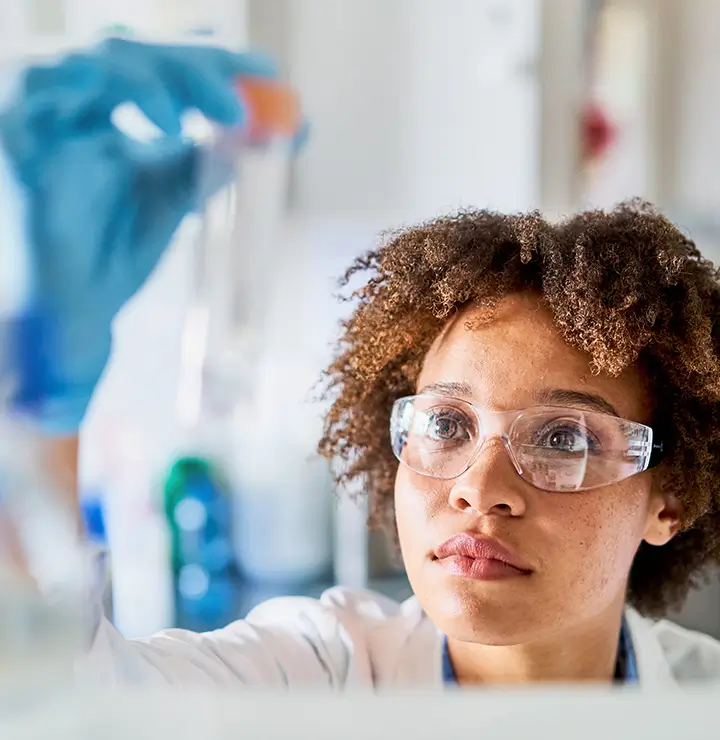 A scientist with goggles and gloves examines a test tube, focused on an experiment in a lab.