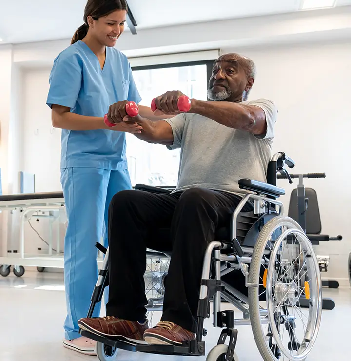 A healthcare worker in scrubs helps an elderly man with dumbbell exercises in a rehab center.