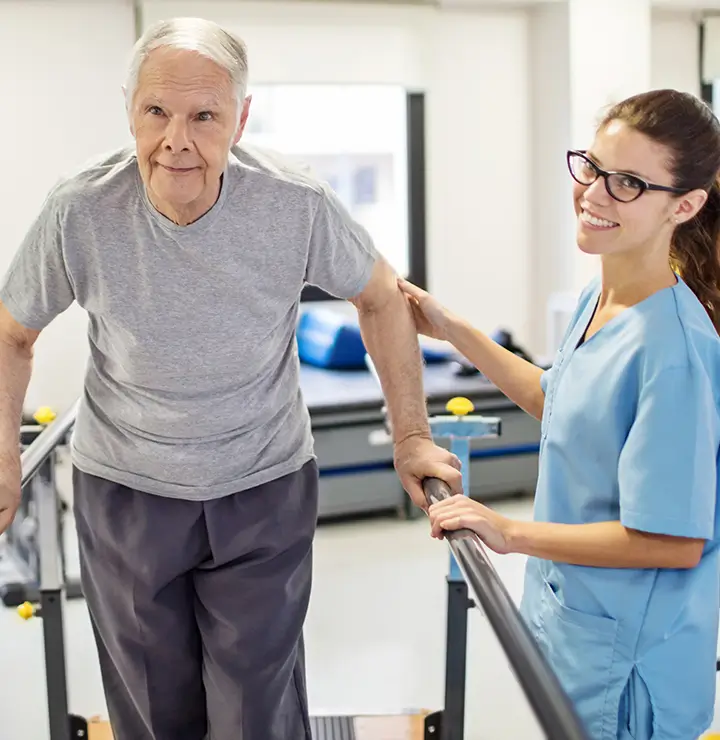 An elderly man walks with support from a smiling healthcare worker in scrubs in a bright rehab room.