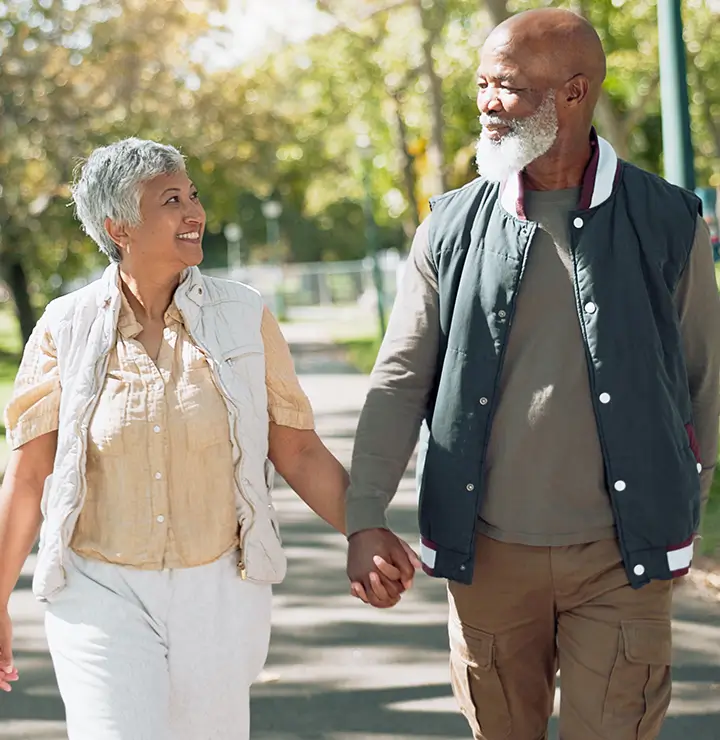 An older couple smiles while walking hand in hand through a sunny park with green trees.