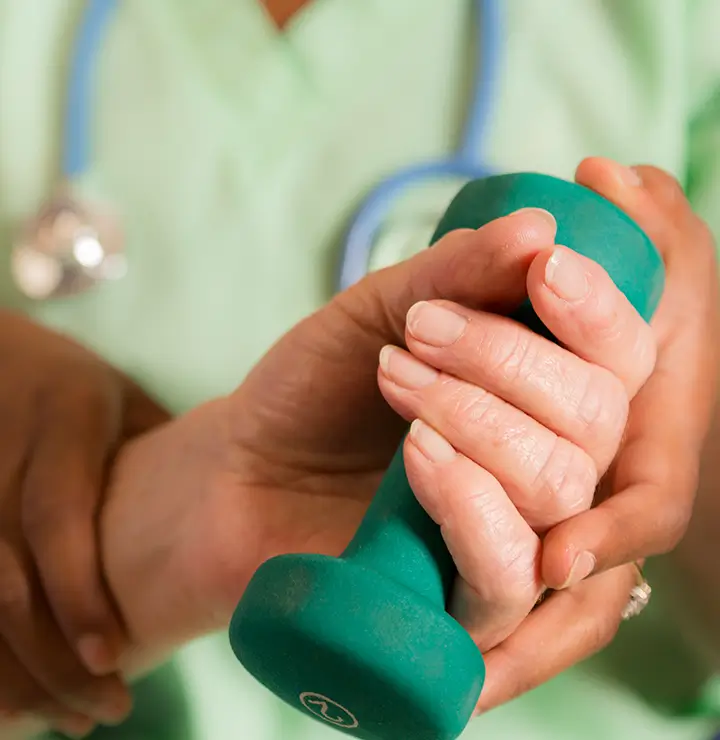 A hand holding a green dumbbell is assisted by another, with a stethoscope in the background.