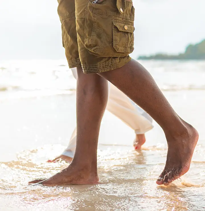 Two people walk barefoot on a sandy beach, waves washing over their feet under an overcast sky.