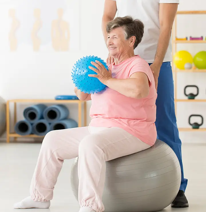 An elderly woman sits on an exercise ball, holding a spiky ball, assisted by a person nearby.