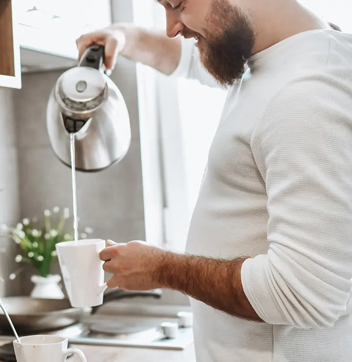 A man pours hot water into a mug in a bright kitchen with a stove and plant in the background.