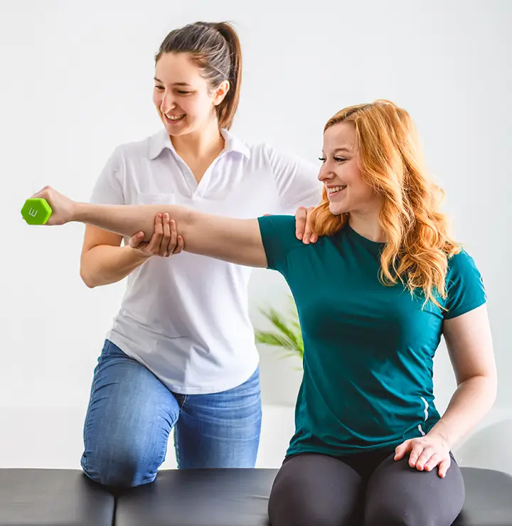 A therapist helps a woman with an arm exercise, holding a dumbbell in a bright, simple room.