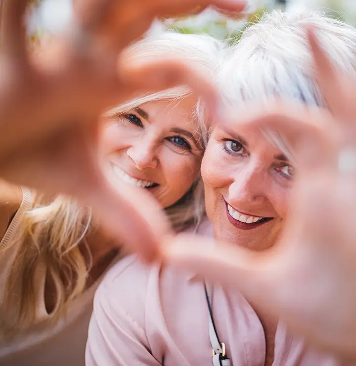 Two smiling women form a heart shape with their hands, focusing on their faces. Close-up shot with a blurred background.