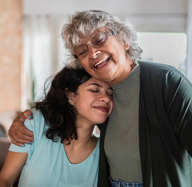 An elderly woman with gray hair hugs a smiling young woman indoors, both expressing warmth and affection.