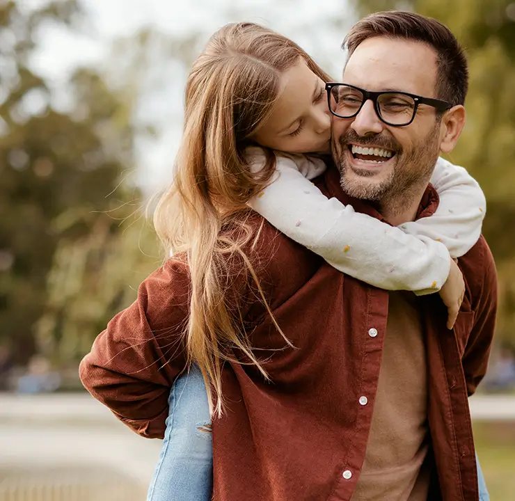 A man wearing glasses smiles as a young girl joyfully hugs him from behind in a park setting.